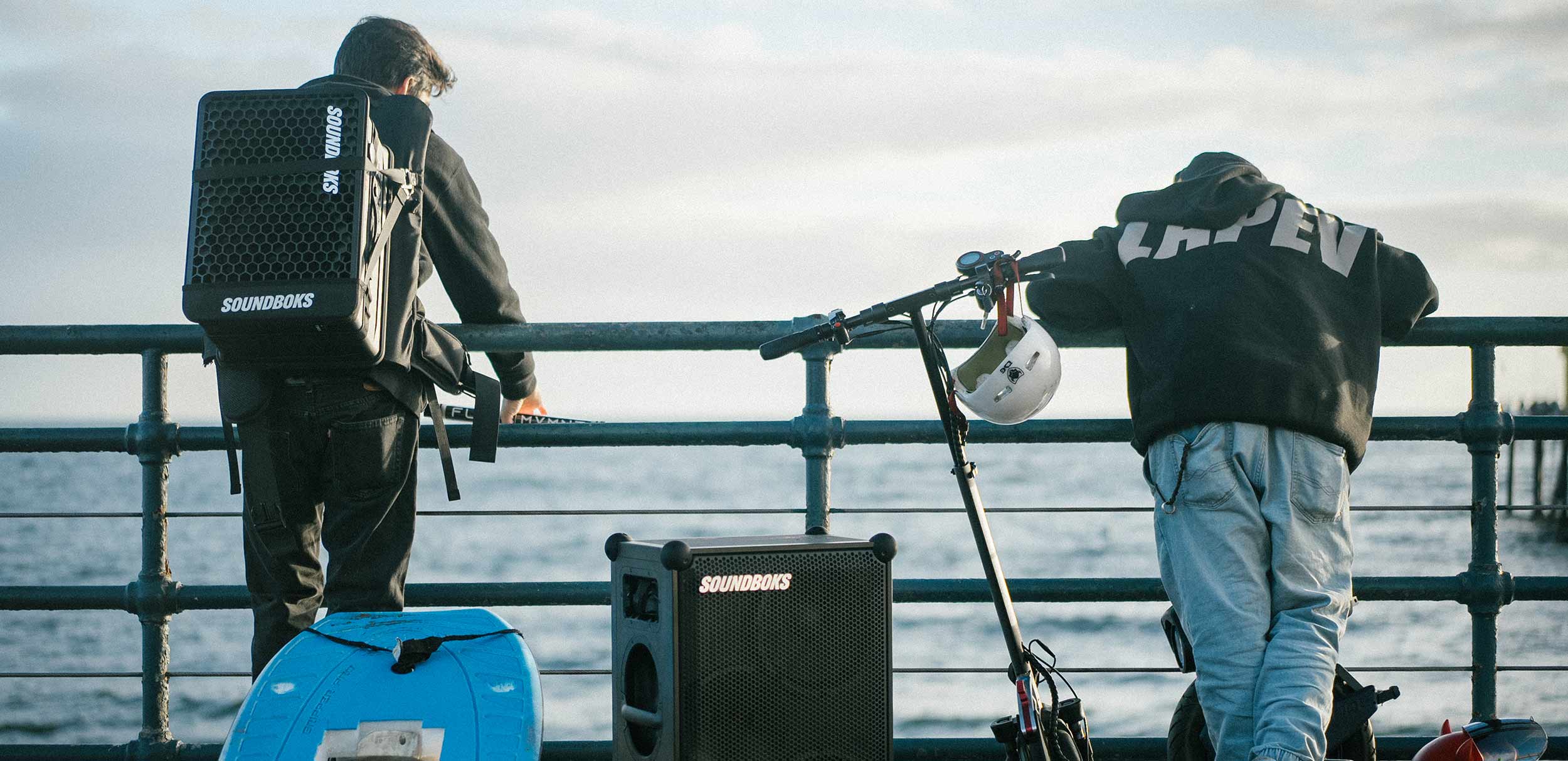 Two persons hanging out at the water with a SOUNDBOKS Go strapped with THE BACKPACK